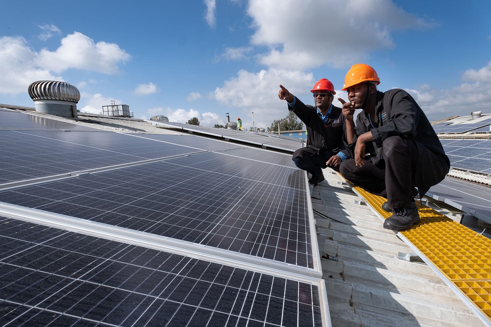 Two men in hard hats kneel by a solar array, pointing towards something in the distance.